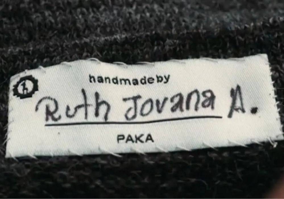 Close-up of garment label showing handwritten ‘Ruth Jovana A.’ under ‘handmade by’ with printed ‘PAKA’ on dark knit background