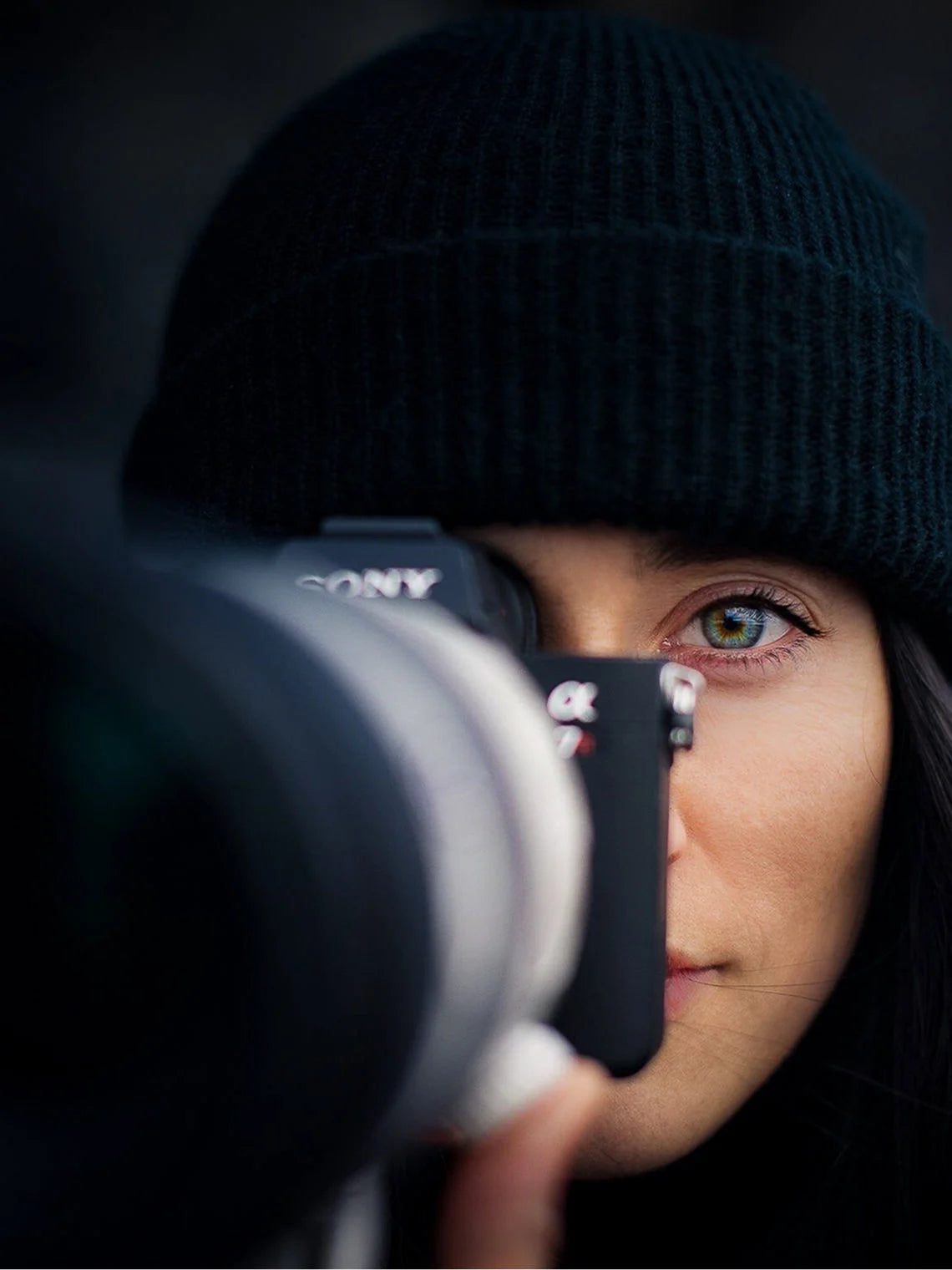 Close-up portrait of a photographer wearing a black knit beanie, aiming a mirrorless camera with the lens in the foreground