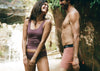 Two models standing on rocks by a waterfall wearing swimwear, woman wearing the Essential Scoop Bralette and Mid-Rise Breif and man wearing the Essential Boxer Brief in Mauve