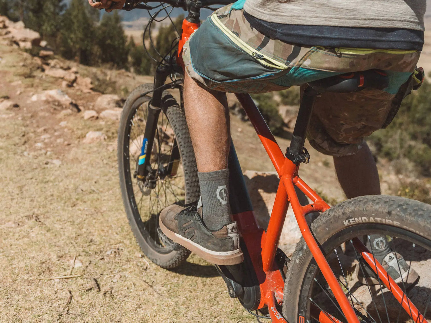 Mountain biker pedaling on a rocky dirt trail wearing gray three-quarter crew socks and brown trail shoes