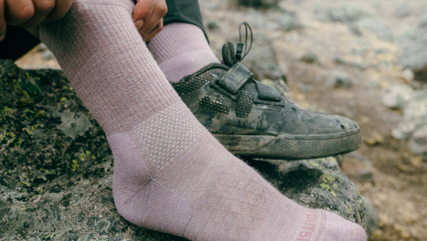 Light beige trail crew sock on a hiker’s foot resting on rocky ground, showing ribbed calf grip and ankle padding