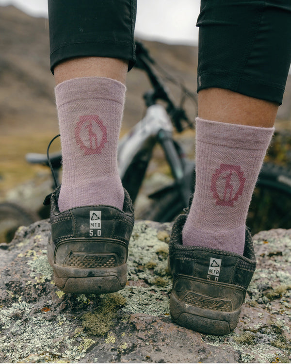 Back view of Trail 3/4 Crew socks in Quail showing mid-calf fit and logo on a model standing on rocky trail with bike