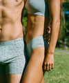Two models wearing light gray alpaca-blend underwear, boxer briefs and high-cut briefs, standing outdoors among greenery