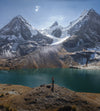Hiker standing on a rocky shore beside a turquoise glacial lake, looking toward snow-capped peaks of Ausangate in Peru