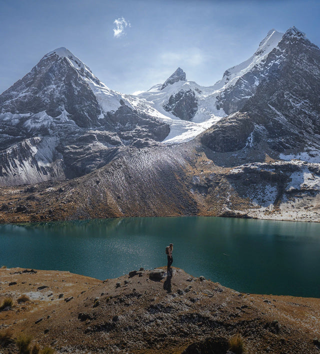 Hiker standing on a rocky shore beside a turquoise glacial lake, looking toward snow-capped peaks of Ausangate in Peru