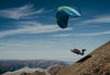 Paraglider flying above a rocky alpine ridge with distant snow-capped mountains under a blue sky