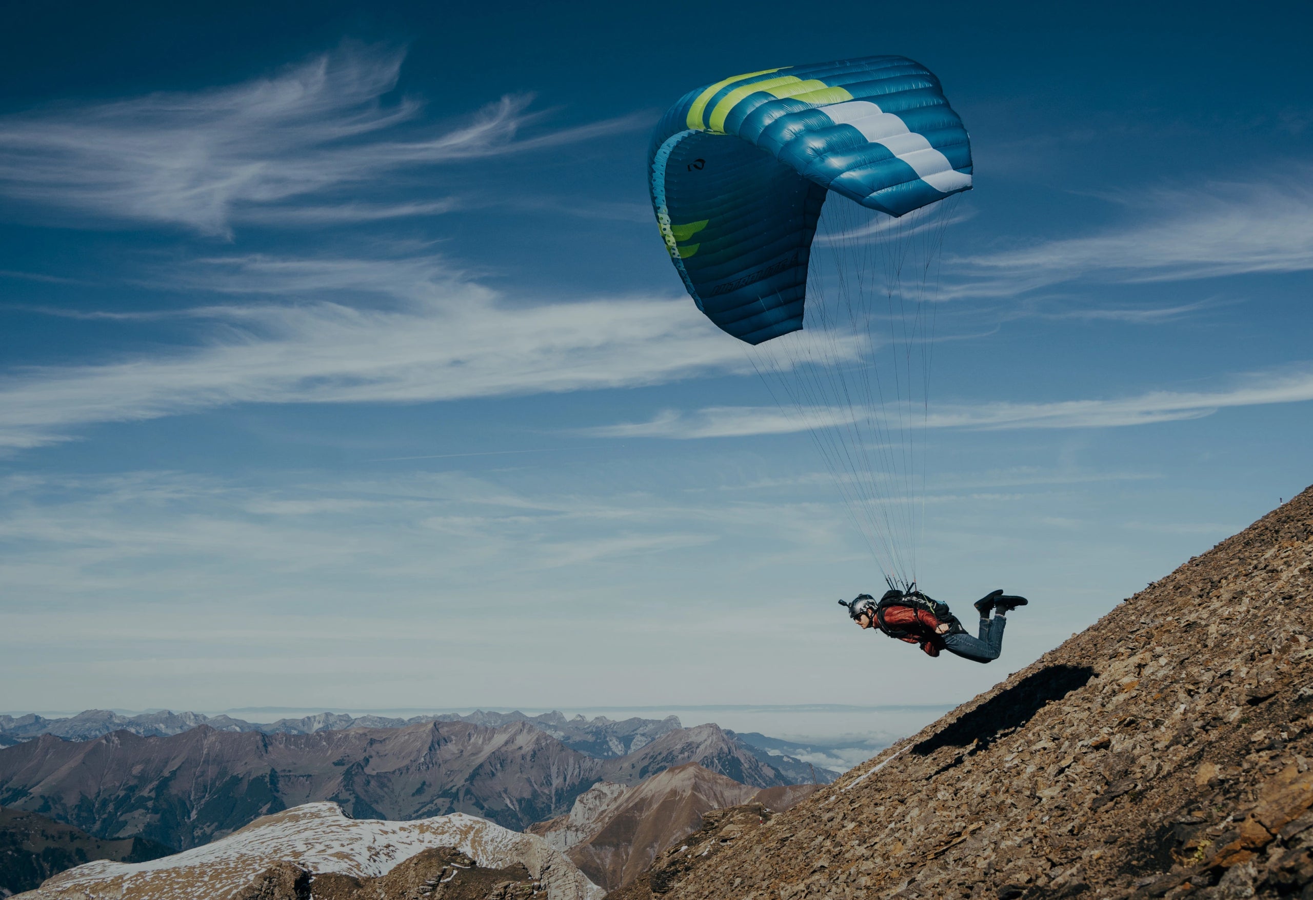 Paraglider flying above a rocky alpine ridge with distant snow-capped mountains under a blue sky