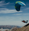 Paraglider soaring above a rugged mountain ridgeline with blue sky and distant peaks