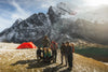Hikers standing by a red tent at an alpine campsite with snow-covered Ausangate peaks in the background