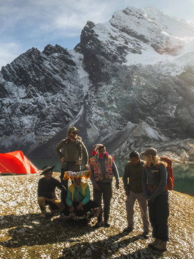 Hikers and local guides standing beside a glacial lake with a red tent and snow-covered mountain peaks