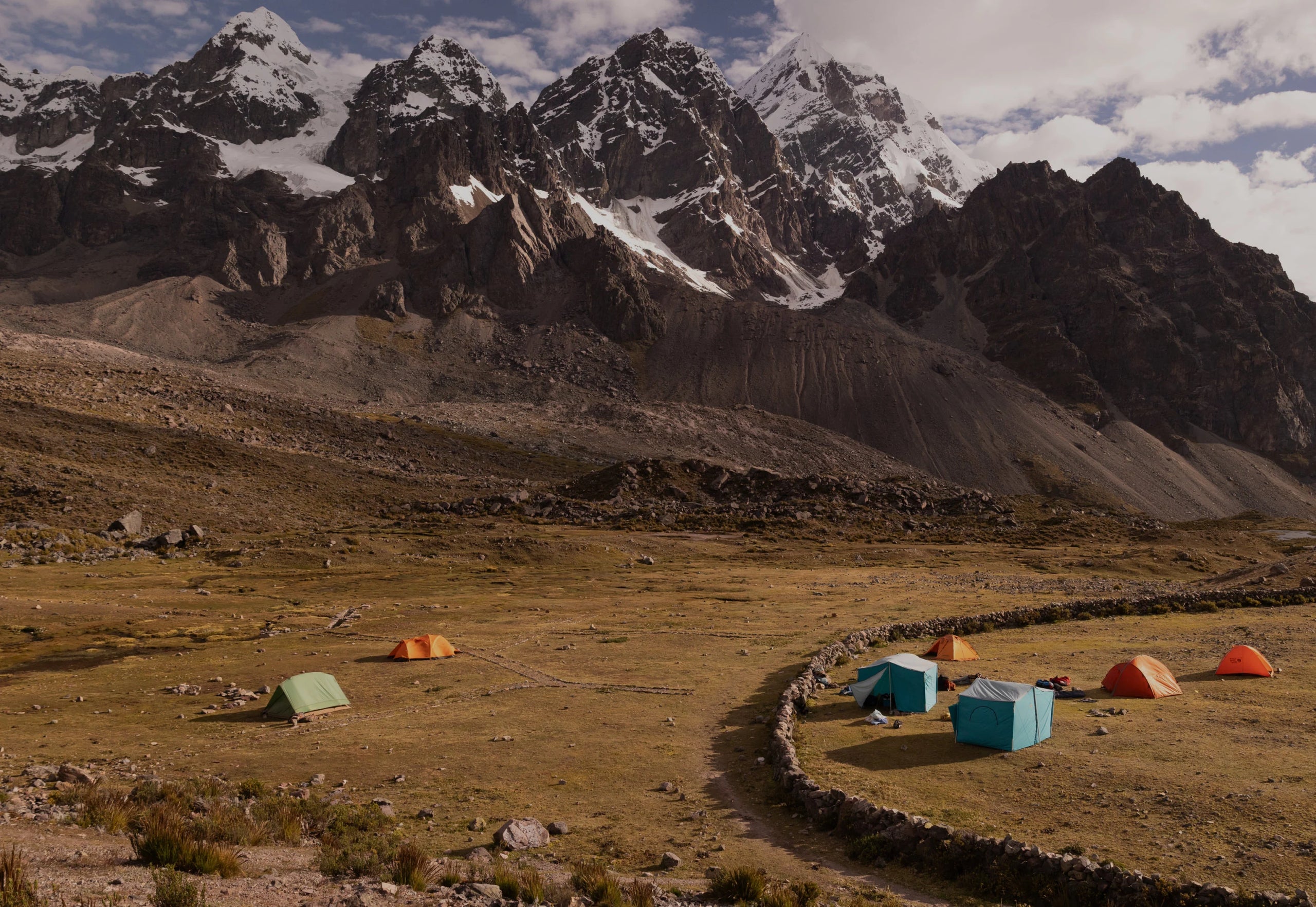 Small campsite with colorful tents pitched on a grassy alpine plain beneath towering snow-capped Ausangate peak