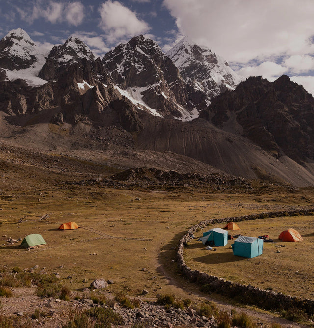 Two tents pitched at a high-altitude campsite beneath snow-capped Ausangate peaks with an overcast sky
