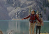 Hiker standing on a mountain lakeshore wearing an orange jacket with alpine peaks reflected in the water