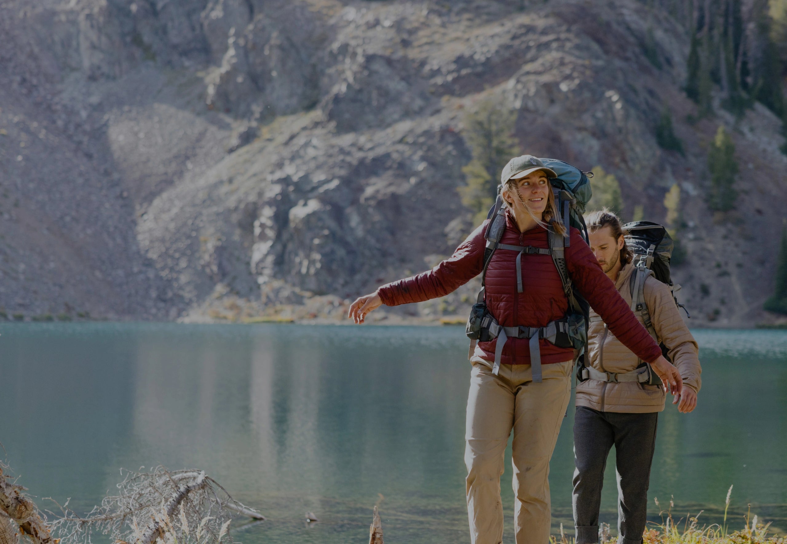 Hiker standing on a mountain lakeshore wearing an orange jacket with alpine peaks reflected in the water