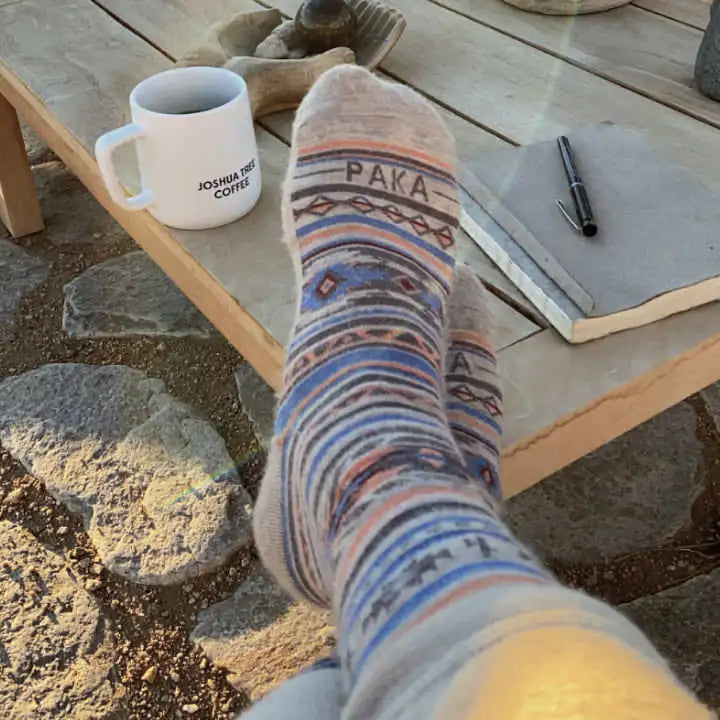 Angled view of Inca Crew socks on crossed feet showing multicolor pattern and PAKA logo on toes beside coffee mug outdoors