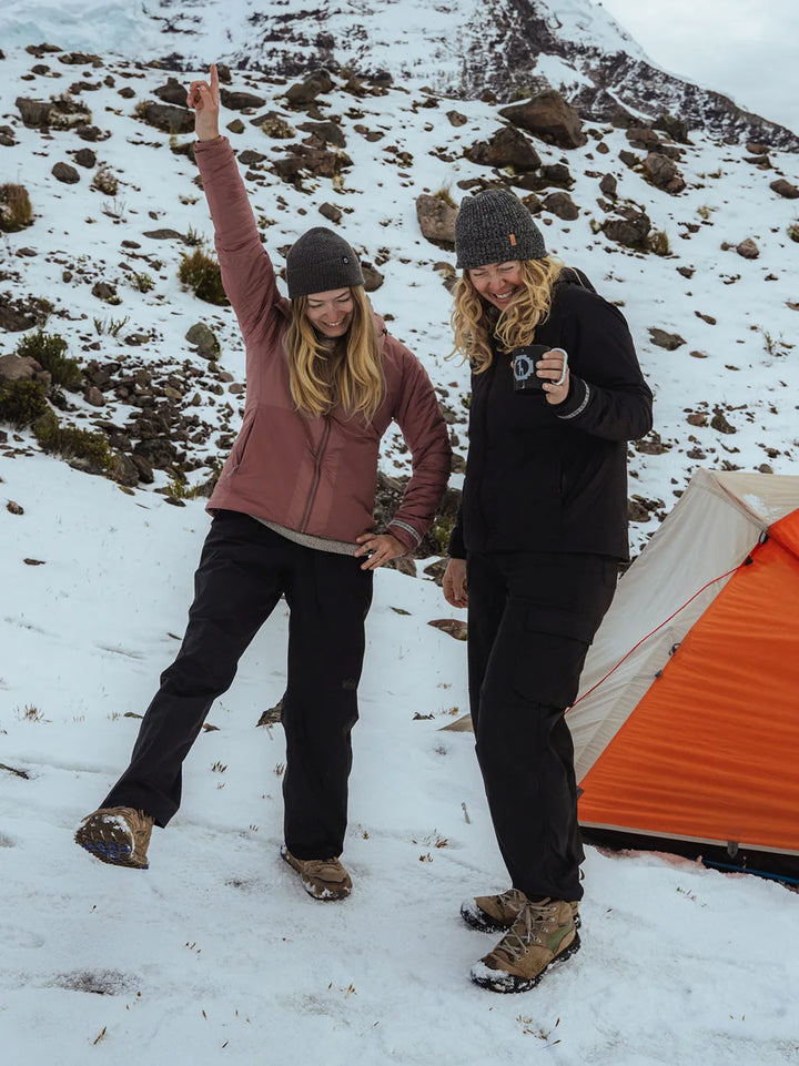 Two hikers standing on snowy ground near an orange tent, one raising an arm, wearing insulated jackets and boots