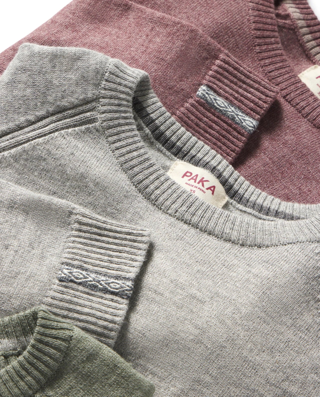 Flat lay of Men’s Ultralight 1.0 Crew showing ribbed crewneck, patterned cuff detail, and PAKA label with gray, maroon, and green sweaters