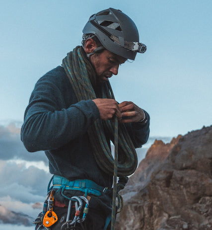 Climber adjusting rope on a rocky slope wearing a dark ultralight crew midlayer and helmet at dawn
