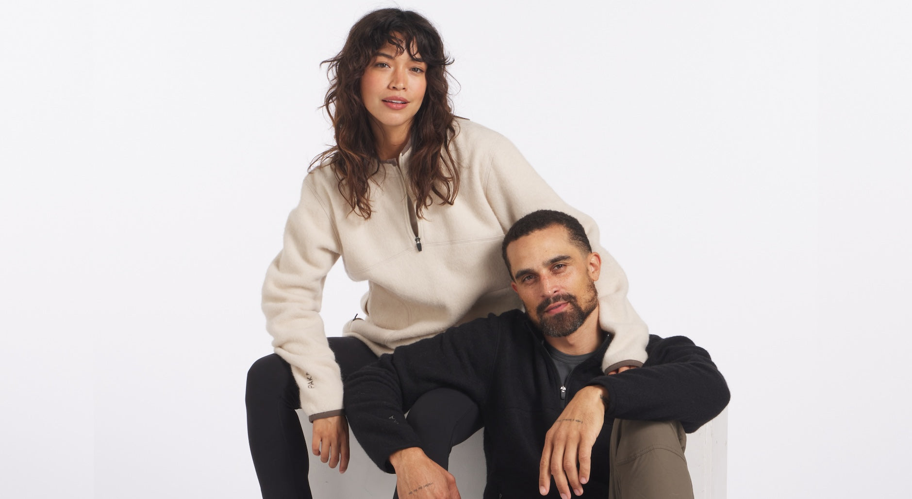 Two models wearing PAKACLOUD alpaca fleece pullovers seated on a light background showing soft brushed texture and relaxed fit