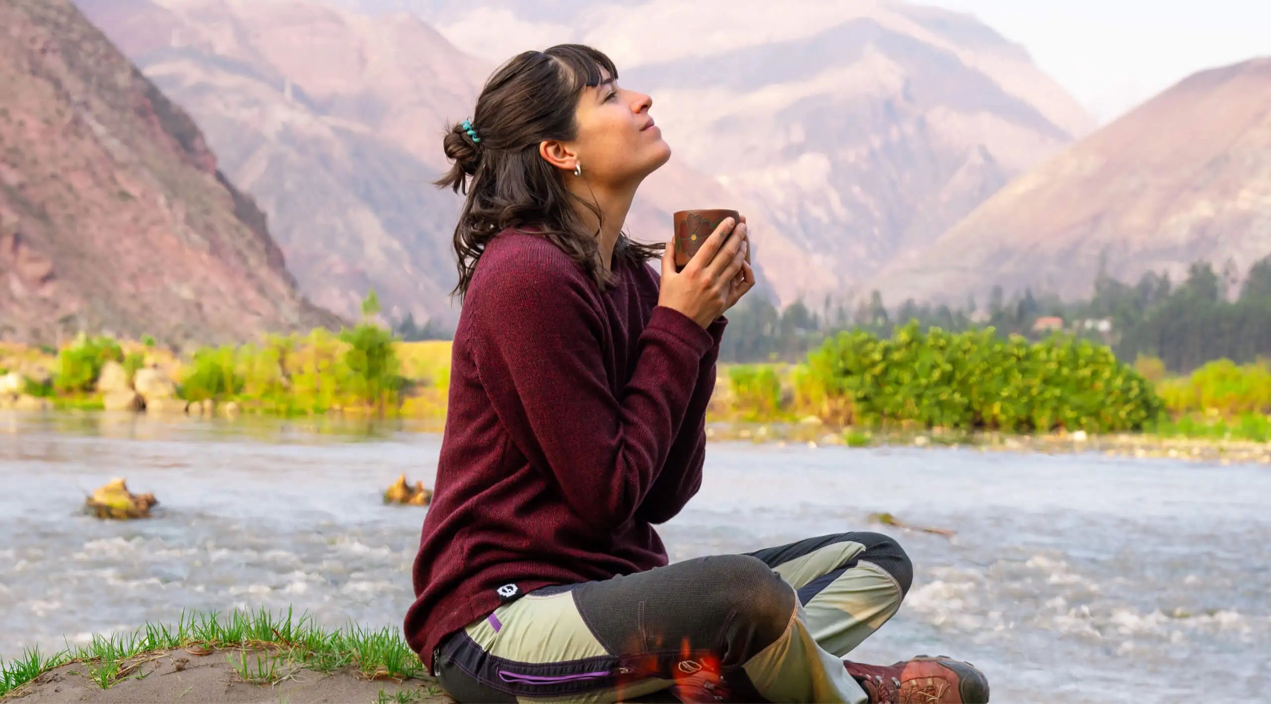 A women wearing a Cusco sweater, holding a cup in front of a river 