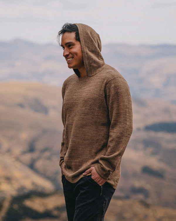 Three-quarter left view of The Hoodie alpaca wool hoodie in Cafe Heather showing relaxed unisex fit and hood against a blurred mountain landscape