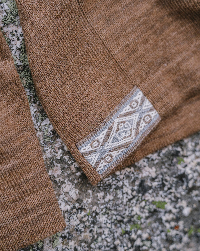 Close up of The Original Crew sweater hem showing cafe heather knit texture and woven geometric tag on a lichen covered rock background