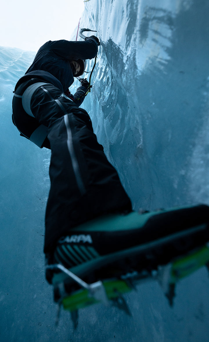Climber ice climbing a vertical glacier wall seen from below, wearing a dark jacket and mountaineering crampons