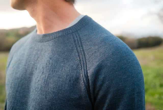 Close-up of men’s blue ultralight alpaca crew sweater showing raglan shoulder and fine knit with blurred outdoor background