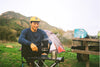 Man wearing a lightweight sweater and hat sitting at a picnic table in a grassy mountain campsite