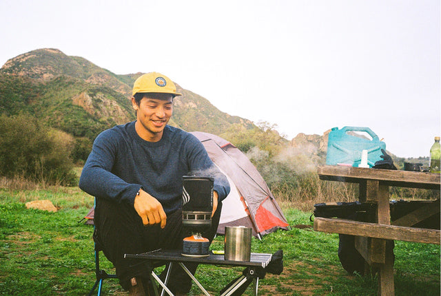 Man wearing a lightweight sweater and hat sitting at a picnic table in a grassy mountain campsite