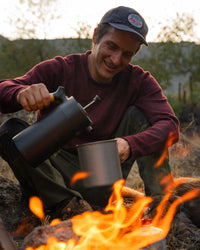Person wearing burgundy The Cusco alpaca merino sweater with relaxed fit pouring coffee into a mug by a campfire at dusk