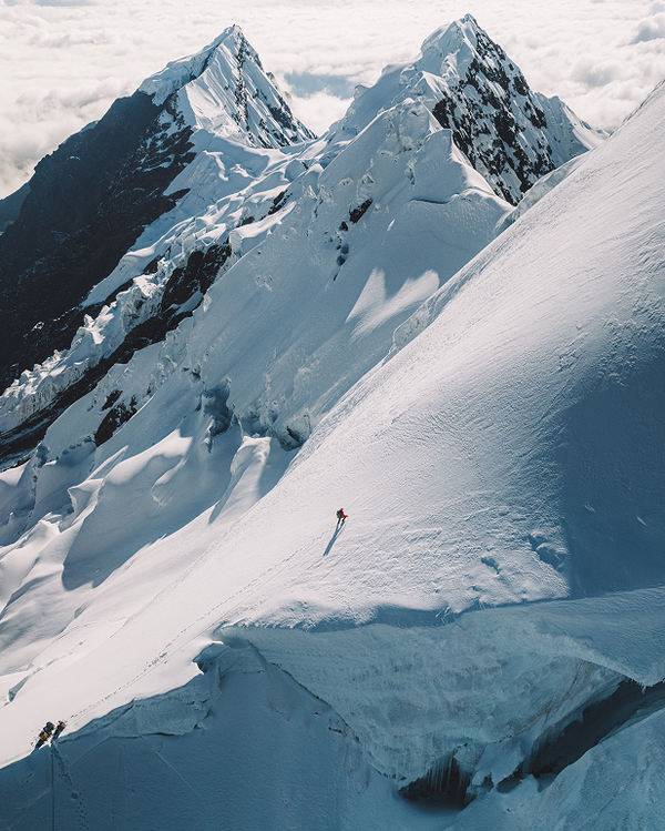 Solo mountaineer in a red jacket traversing a steep snow ridge beneath jagged alpine peaks and a sea of clouds