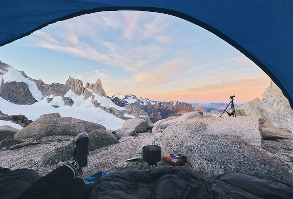 View from inside a tent showing a camper’s legs and feet looking out at alpine mountains at sunrise with camping gear on the rocky foreground
