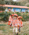 Children in orange traditional Peruvian ponchos and hats walking on a grassy hillside with a blue-roofed building