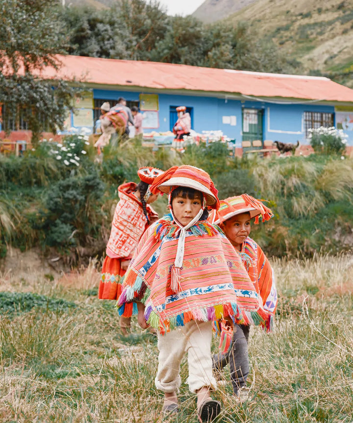 Children in orange traditional Peruvian ponchos and hats walking on a grassy hillside with a blue-roofed building