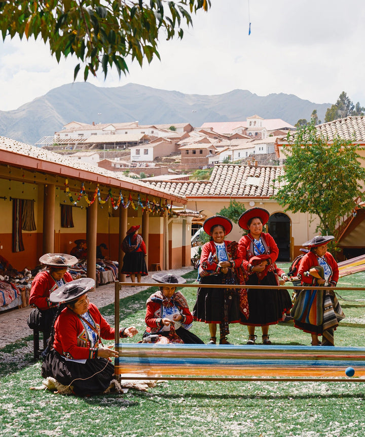 Quechua weavers in colorful traditional dress seated in a courtyard with handmade textiles and Andes mountains in the background