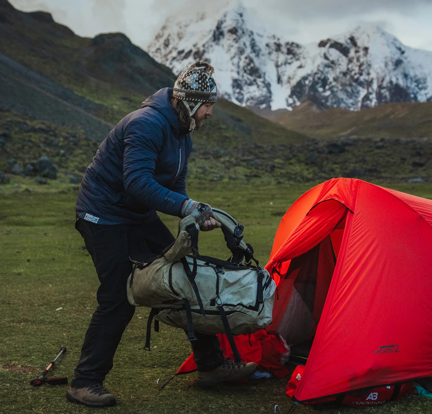 Hiker loading gear into a red tent at a mountain campsite with snow-covered peaks in the background