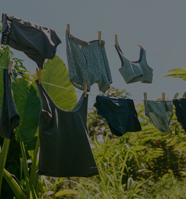 PAKA Essentials underwear and tops hanging on a clothesline in a sunlit grassy outdoor setting