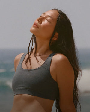 Woman wearing a fitted dark gray scoop-neck bra facing upward with eyes closed on a sunlit beach