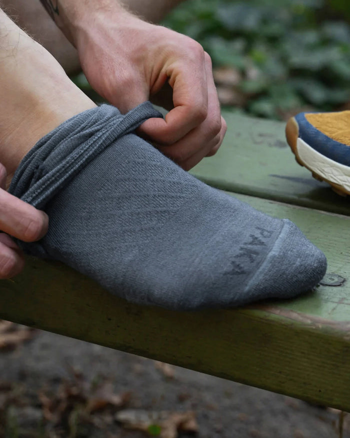 Close-up of Essential Crew Sock in indigo being pulled onto a foot, showing PAKA branding and ribbed cuff on an outdoor bench