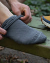 Close-up of Essential Crew Sock in indigo being pulled onto a foot, showing PAKA branding and ribbed cuff on an outdoor bench