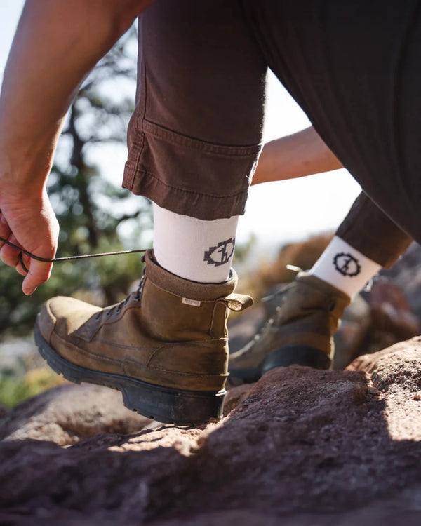 Close-up side view of Essential Crew Socks in white showcasing crew height and cuff logo worn with hiking boots on a rocky trail