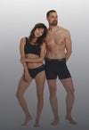 Front view of couple modeling black PAKA alpaca underwear: slim-fit men’s boxer briefs and women’s brief on gray background