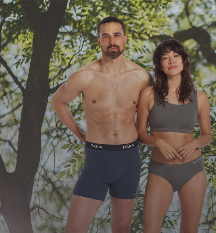 Front view of a man in navy PAKA alpaca boxer briefs and a woman in gray alpaca briefs showing PAKA waistband and athletic fit with trees behind