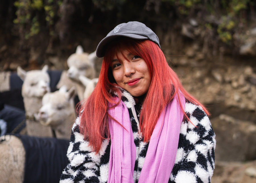 Person with red hair and a gray cap standing among llamas outdoors