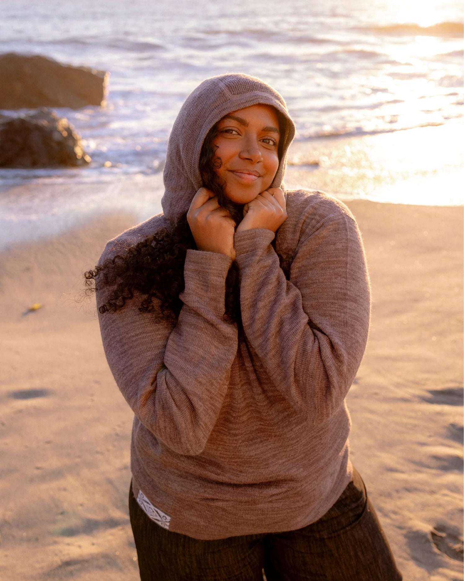 Woman Named Meghna in The Hoodie Flint Sweater Hoodie pulling her hood close on a beach with water behind her – PAKA Apparel