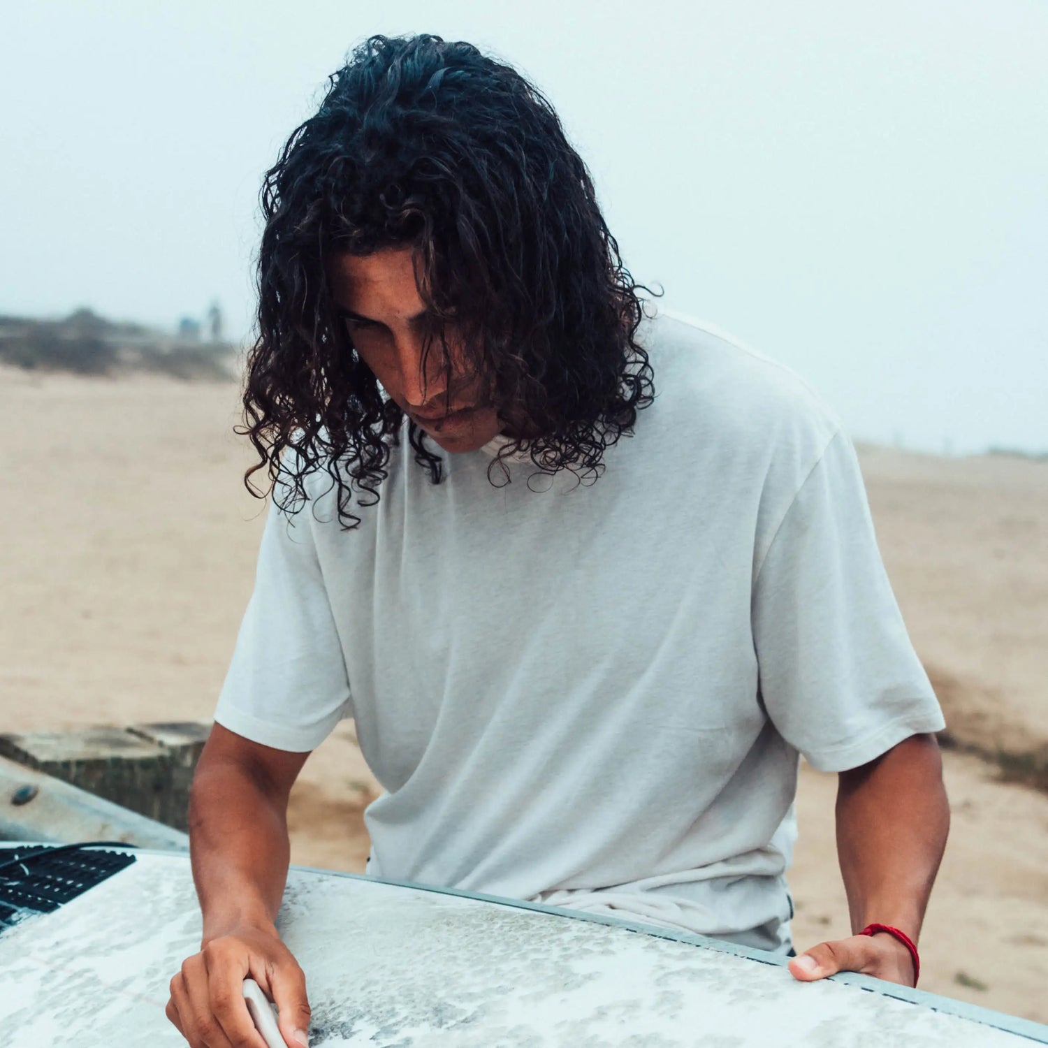 Man with Long, Wavy Hair in Cream / White Alpaca T-Shirt Waxing His Surfboard on a Beach – PAKA Apparel