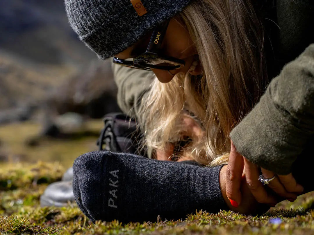Close-up of dark grey ankle sock with PAKA logo on a woman’s foot, low-cut fit on grass in an outdoor mountain setting