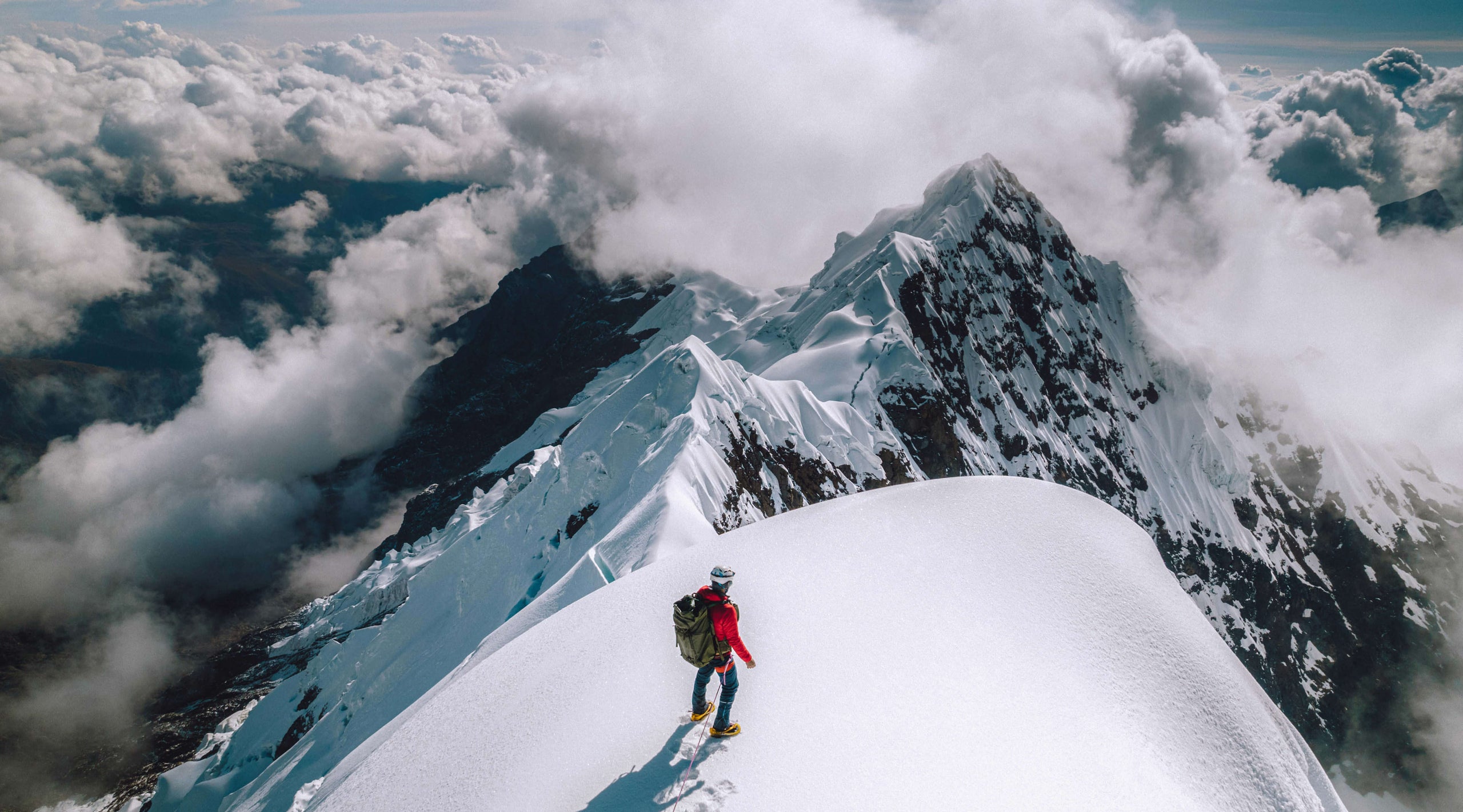 Two climbers traversing a narrow snowy ridge toward a jagged peak in the Cordillera Blanca with clouds overhead