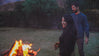 Two people standing by a campfire at dusk in a wooded outdoor setting